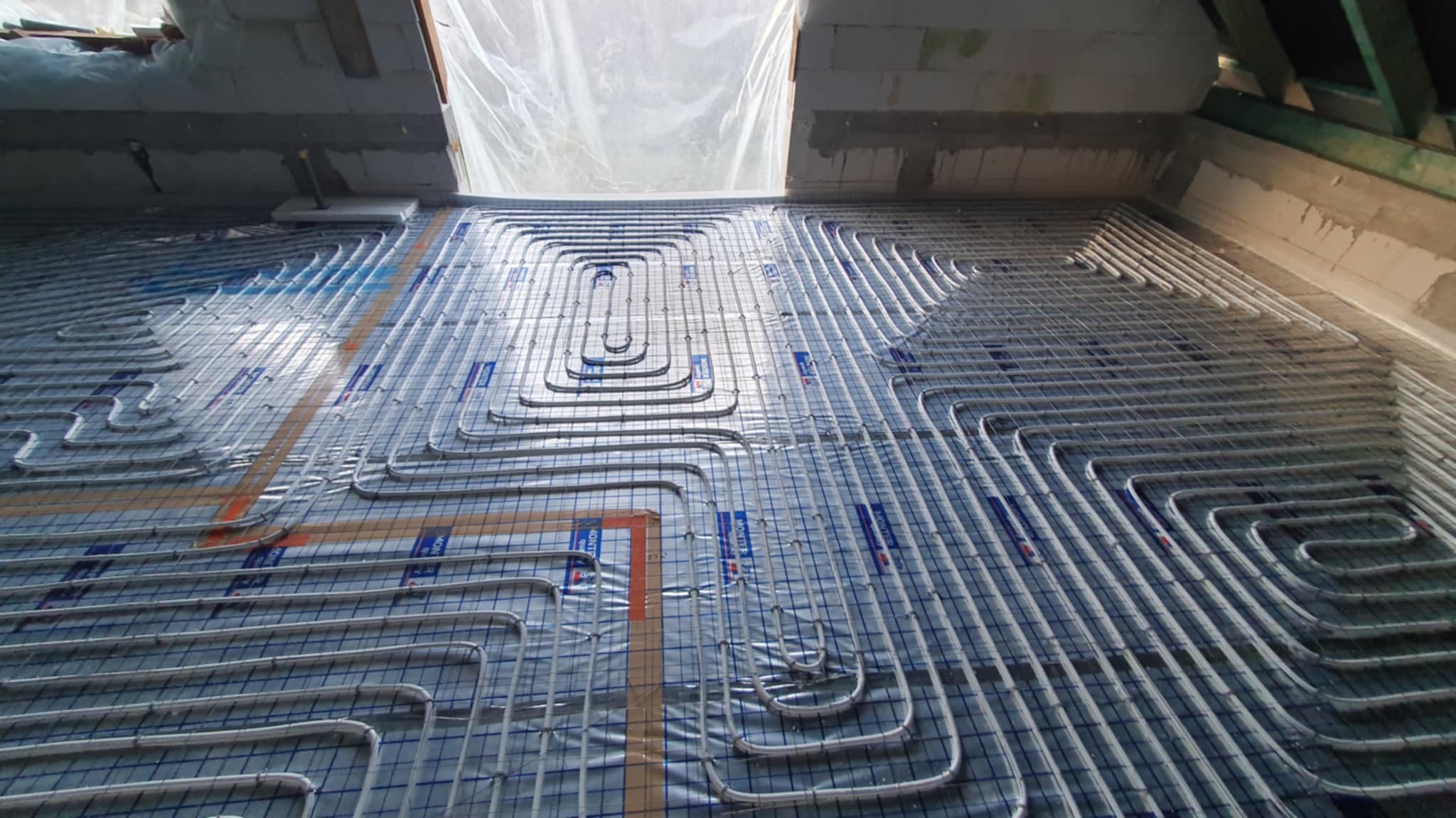 Rozłożona instalacja ogrzewania podłogowego z rurami grzewczymi ułożonymi w pętle na folii z siatką, widok z góry na surowy strop.