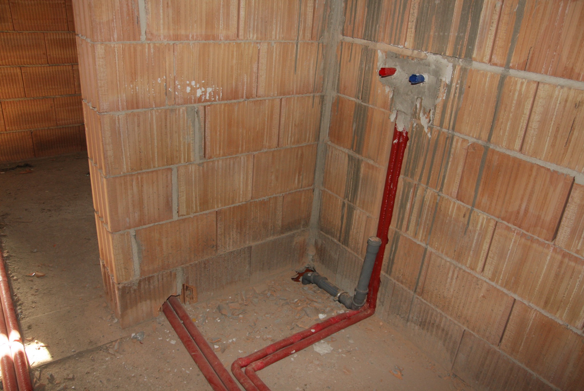 Surowy stan: czerwone rury instalacji wodnej wystające ze ściany z pustaków ceramicznych, z widocznymi zaślepkami ciepłej i zimnej wody, oraz szare rury odpływowe na podłodze w trakcie montażu.