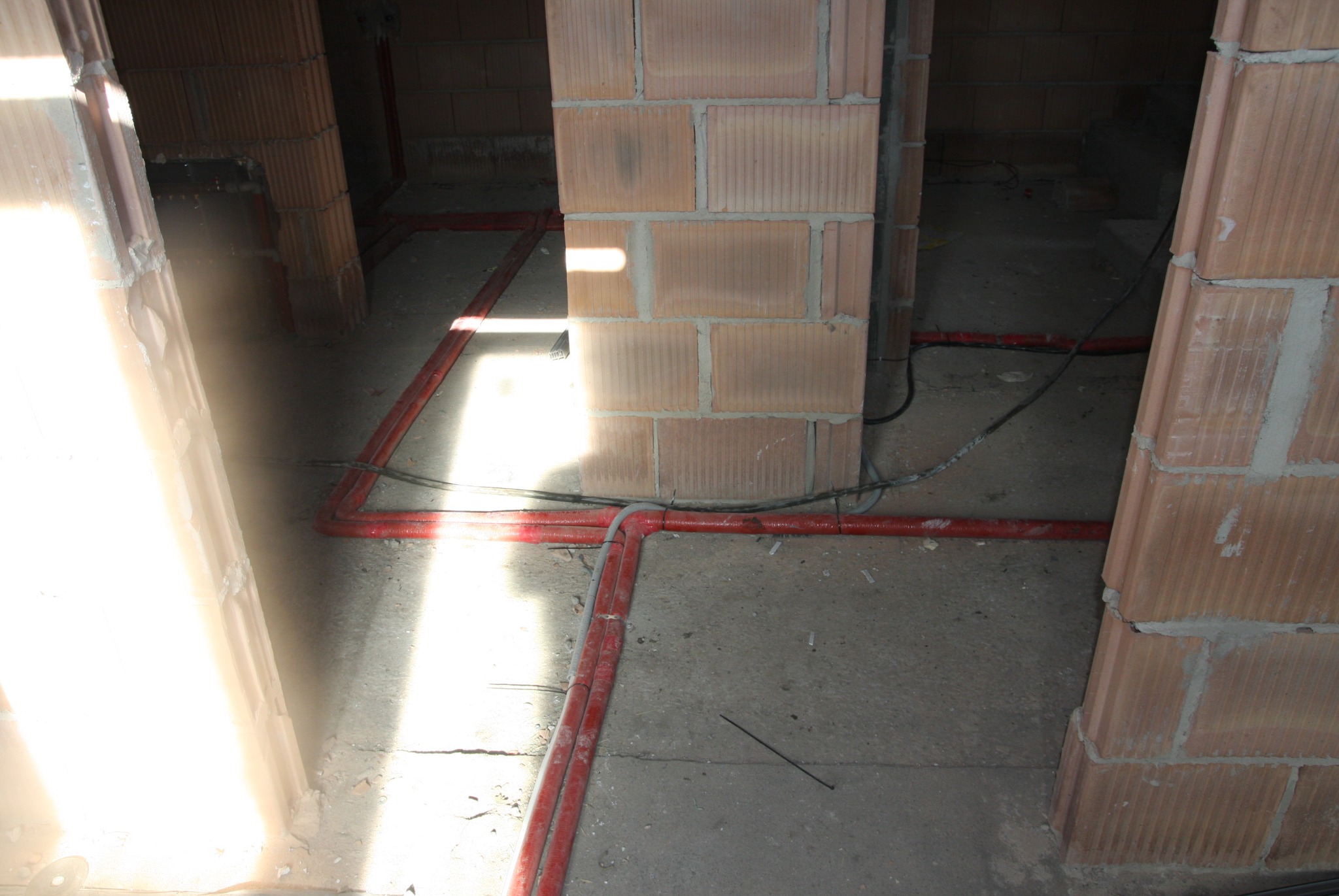 Czerwone rury instalacji grzewczej ułożone na surowym betonowym podłożu w trakcie budowy, widoczne fragmenty ścian z cegły ceramicznej.