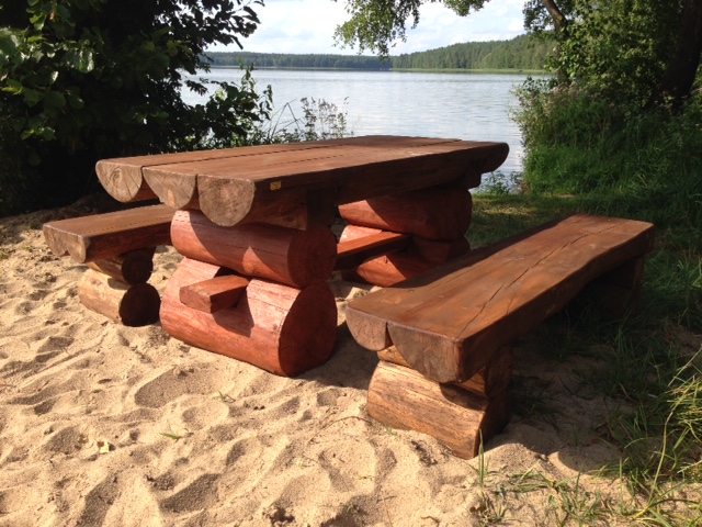 Drewniany stół i ławki z bali na piaszczystym brzegu jeziora, widoczne słoje drewna i naturalne wykończenie.
