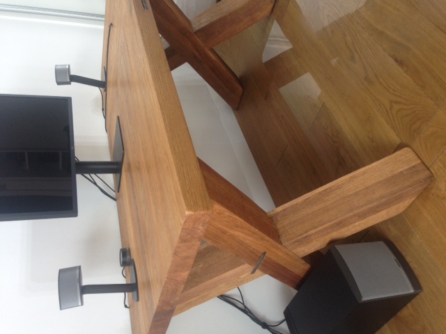 Solidny stół z drewna z krzyżowymi nogami, wyposażony w zamontowany telewizor i głośniki, ustawiony na parkiecie.