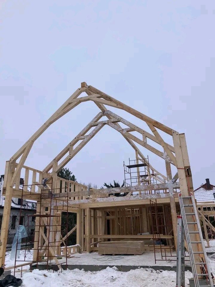 Konstrukcja drewniana domu w trakcie budowy, widoczne rusztowania i drabina, zimowa sceneria z pokrywą śnieżną.