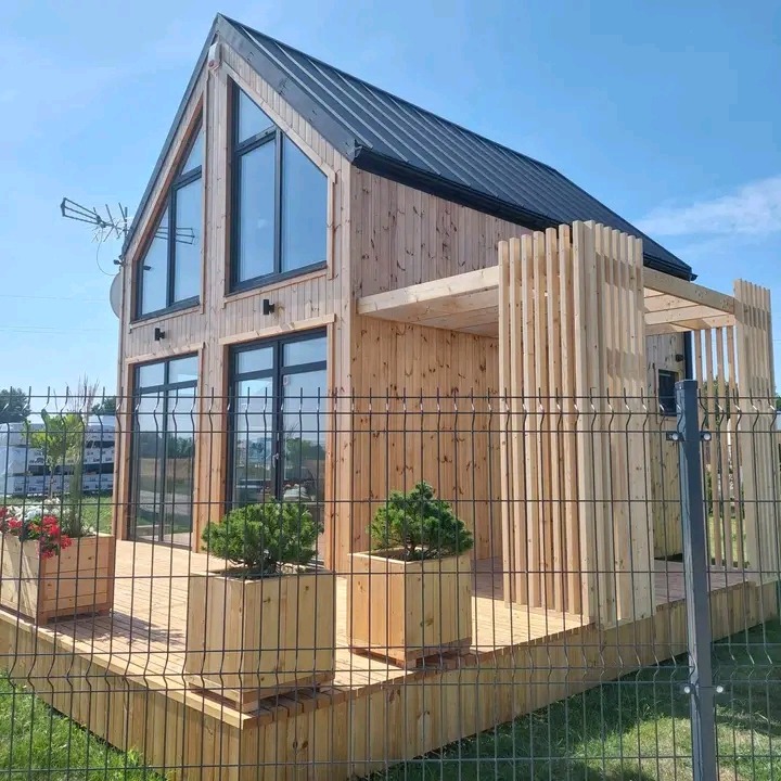 Nowoczesny, parterowy dom z drewna o skośnym dachu i dużych oknach, otoczony drewnianym tarasem z donicami i ogrodzeniem z siatki.
