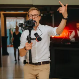 Mężczyzna w okularach, z kamerą Sony Alpha na gimbalu DJI, unosi palec wskazujący w górę podczas filmowania wydarzenia, w tle widoczni bawiący się goście.