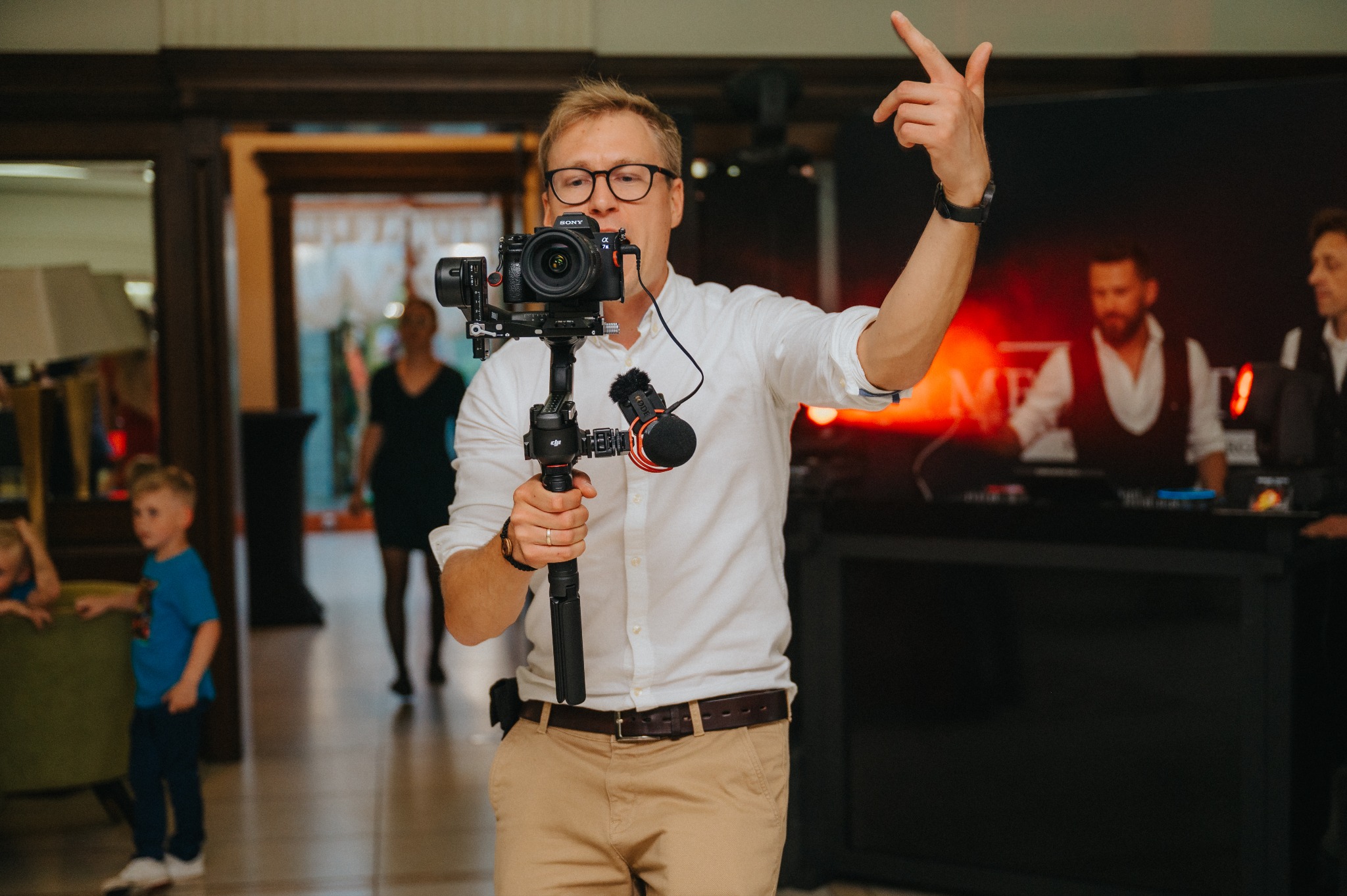 Mężczyzna w okularach, z kamerą Sony Alpha na gimbalu DJI, unosi palec wskazujący w górę podczas filmowania wydarzenia, w tle widoczni bawiący się goście.