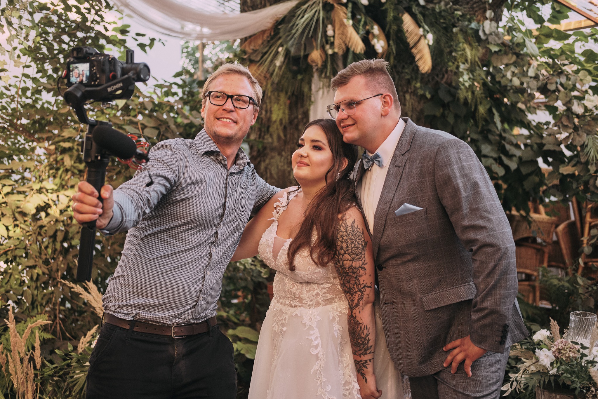 Uśmiechnięty mężczyzna z kamerą na stabilizatorze filmuje parę młodą w otoczeniu dekoracji roślinnych podczas ceremonii ślubnej.
