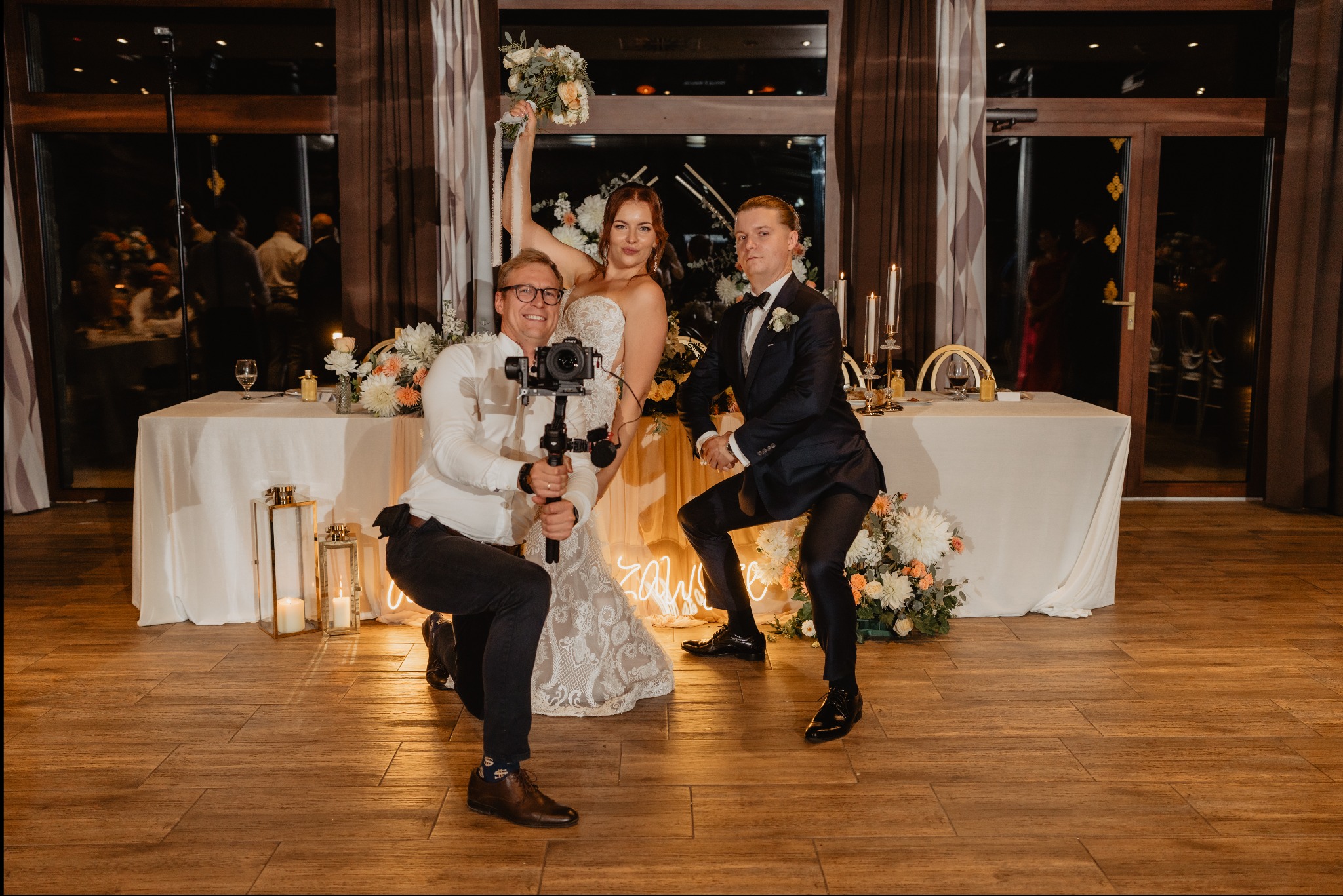 Uśmiechnięta para młoda pozuje z kamerzystą trzymającym stabilizator kamery i gościem w garniturze, na drewnianej podłodze, w tle stół z dekoracjami kwiatowymi i świecami.