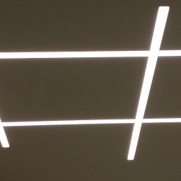 PRW Adm Pikul - Nowoczesne oświetlenie sufitowe z czterema przecinającymi się liniami LED, tworzącymi geometryczny wzór kwadratów i prostokątów, na tle beżowego sufitu.