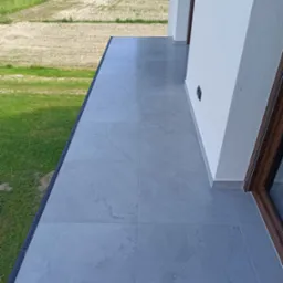 Wyłożony jasnoszarymi płytkami balkon z widokiem na zieleń, widoczna ściana budynku z oknem.