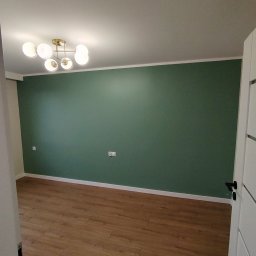 PRW Adm Pikul - Pokój po remoncie: ściana pomalowana na butelkową zieleń, jasnobrązowe panele podłogowe, biała listwa przypodłogowa i sufitowa, nowoczesna lampa sufitowa z mlecznymi kulami.