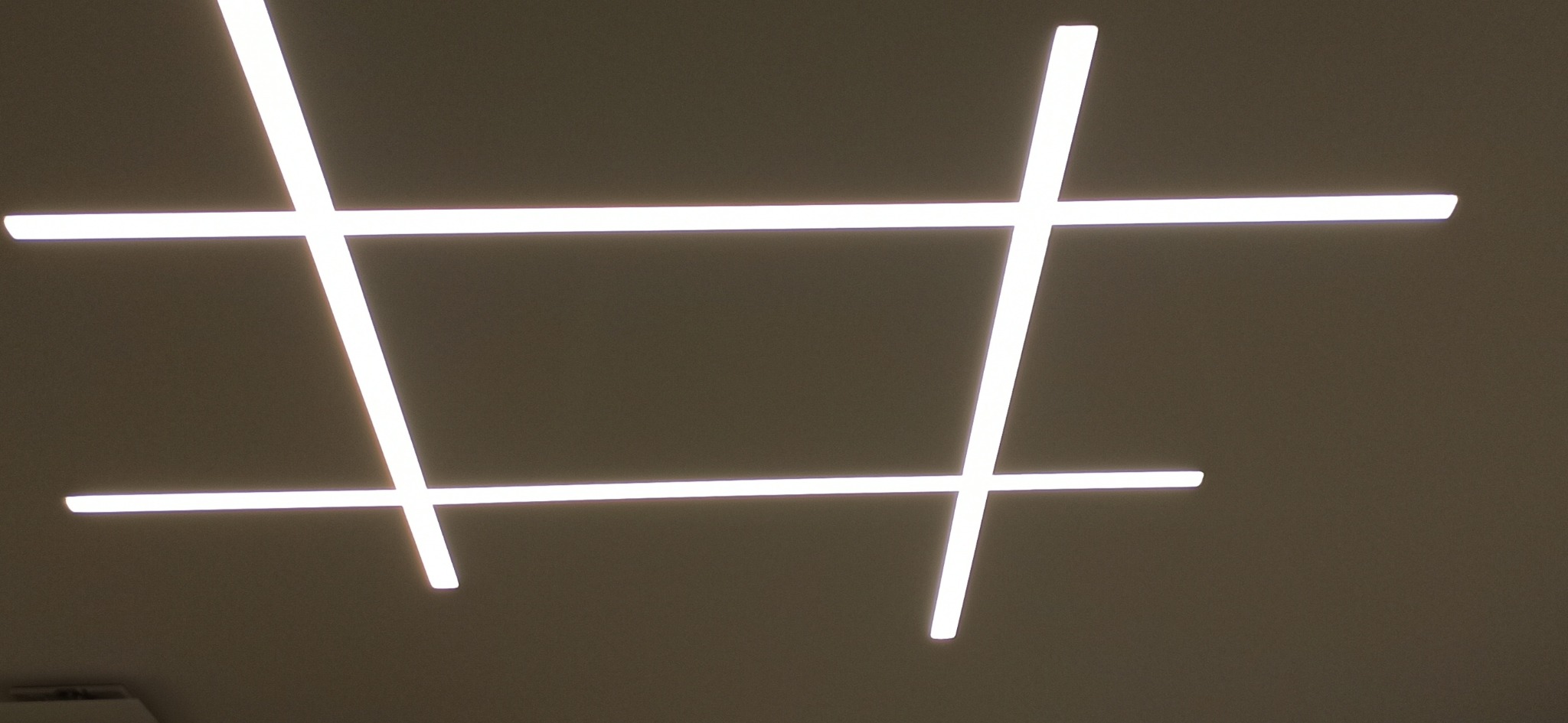 Nowoczesne oświetlenie sufitowe z czterema przecinającymi się liniami LED, tworzącymi geometryczny wzór kwadratów i prostokątów, na tle beżowego sufitu.
