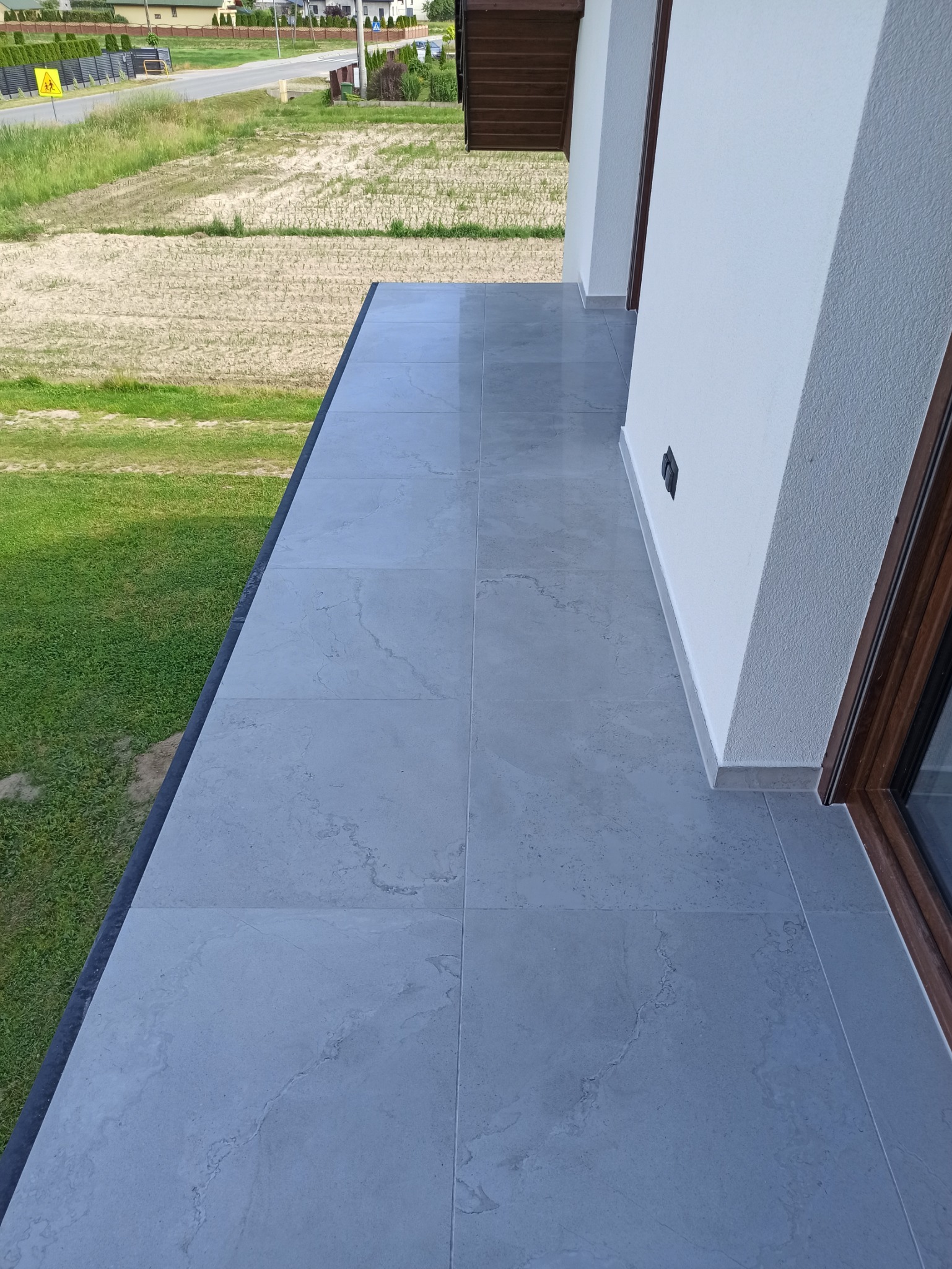 Wyłożony jasnoszarymi płytkami balkon z widokiem na zieleń, widoczna ściana budynku z oknem.