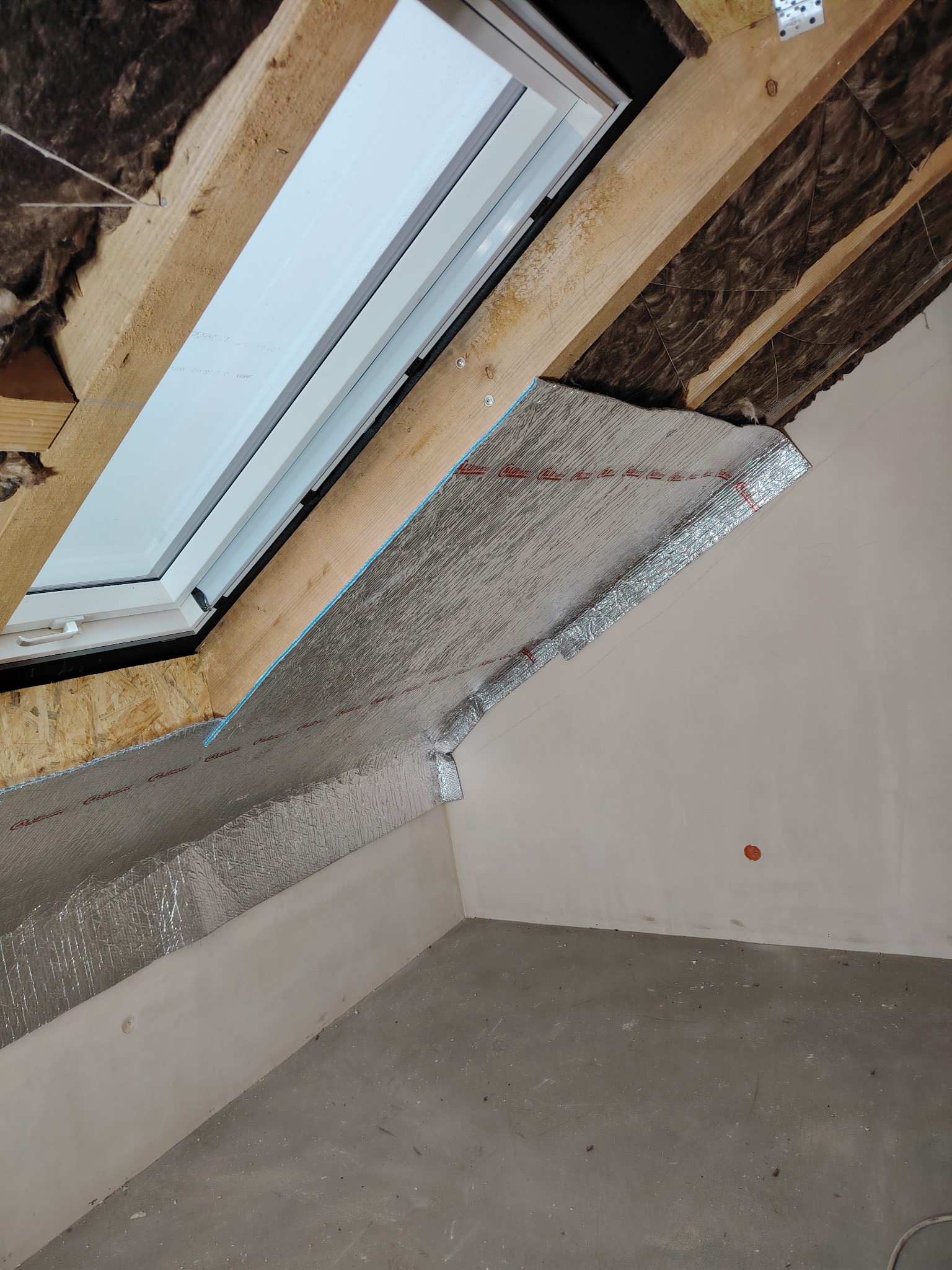 Montaż okna dachowego z izolacją termiczną folią aluminiową na poddaszu, widoczne drewniane elementy konstrukcyjne i wełna mineralna.