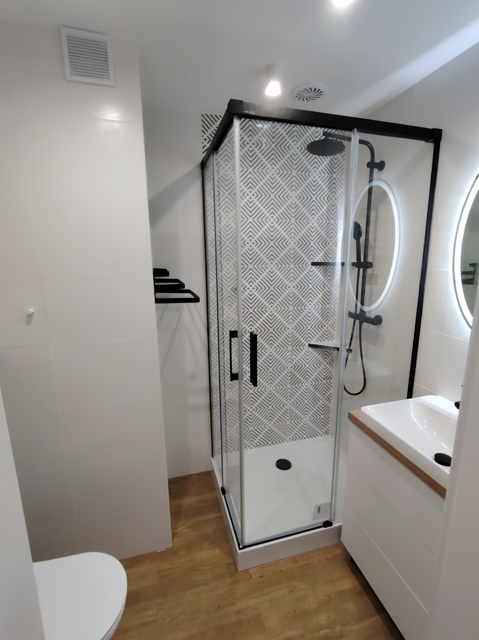 Nowoczesna łazienka z kabiną prysznicową typu walk-in z czarnymi ramami, wzorzystymi płytkami na ścianie wewnątrz kabiny, umywalką nablatową na białej szafce z drewnianym blatem i okrągłym lustrem...