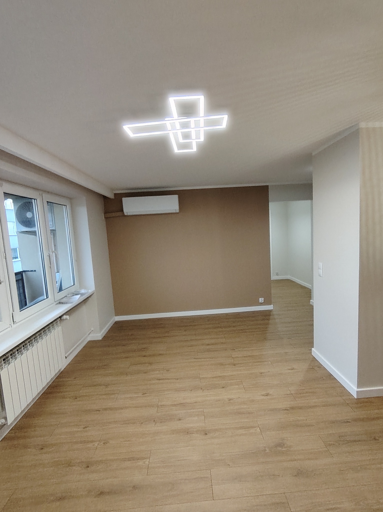 Pusty pokój po remoncie z drewnianą podłogą, brązową ścianą, białymi listwami przypodłogowymi i geometryczną lampą sufitową.