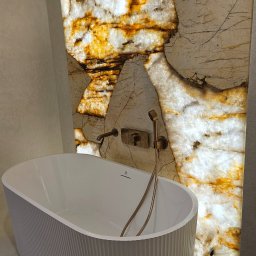 DEKOM - Nowoczesna biała wanna wolnostojąca z ozdobnym żłobkowaniem, na tle podświetlanej ściany z onyksu z armaturą w kolorze szczotkowanego złota.