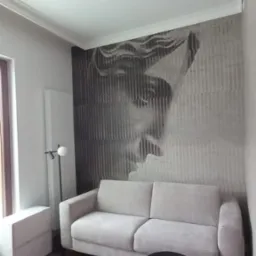 Jasnoszara sofa z dwoma poduszkami na tle ściany z fototapetą przedstawiającą twarz, obok czarny stolik kawowy na metalowych nogach i lampa podłogowa.