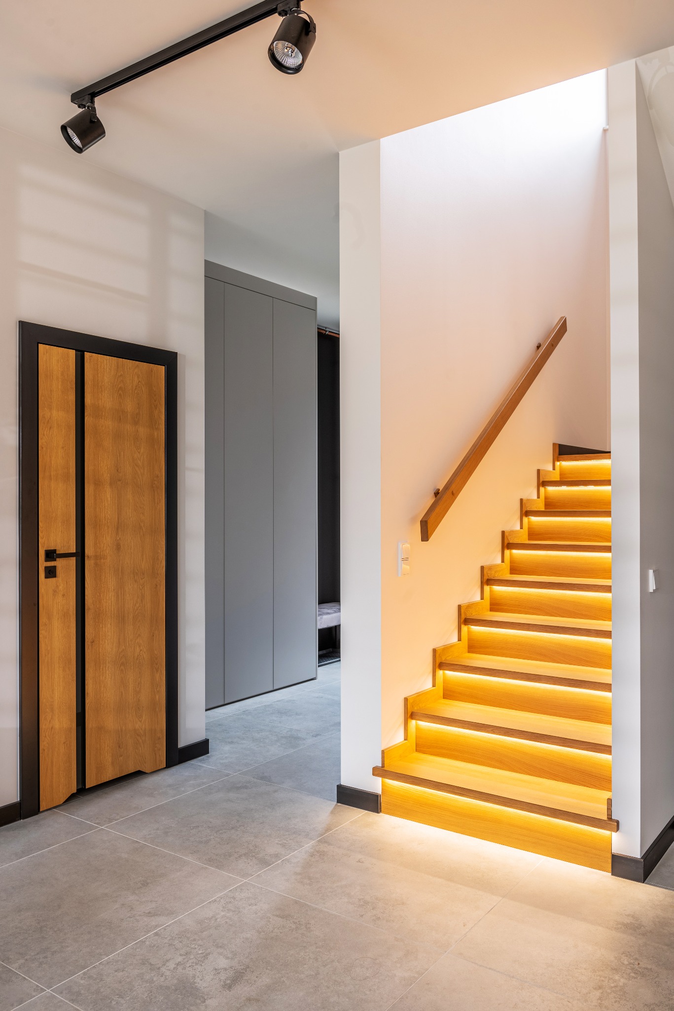 Nowoczesne schody drewniane z podświetleniem LED, drewniane drzwi z czarną ramą i szara szafa wnękowa w minimalistycznym wnętrzu.