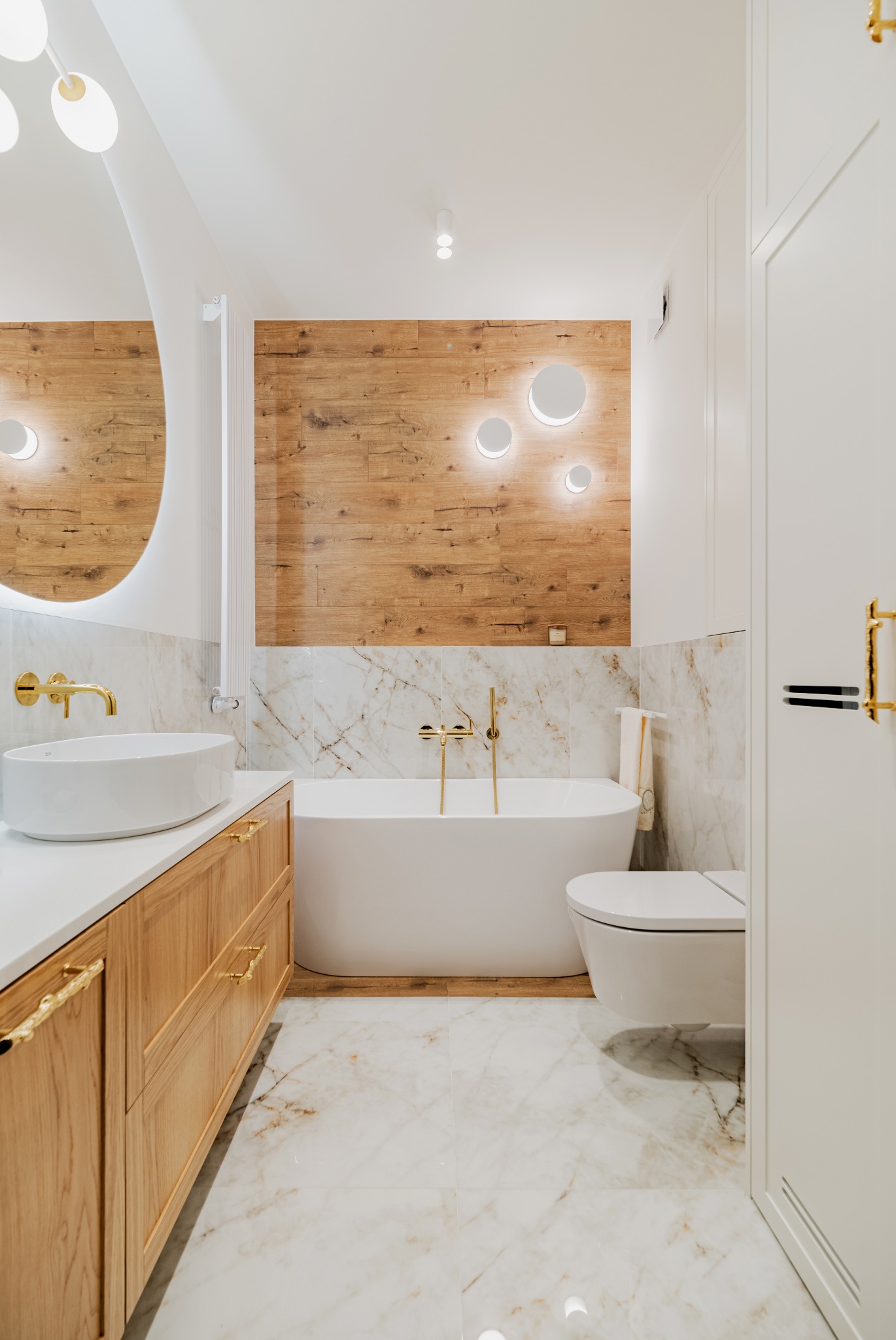 Nowoczesna łazienka z drewnianą ścianą, marmurowymi płytkami, białym blatem i złotą armaturą, oświetlona okrągłymi lampami LED.
