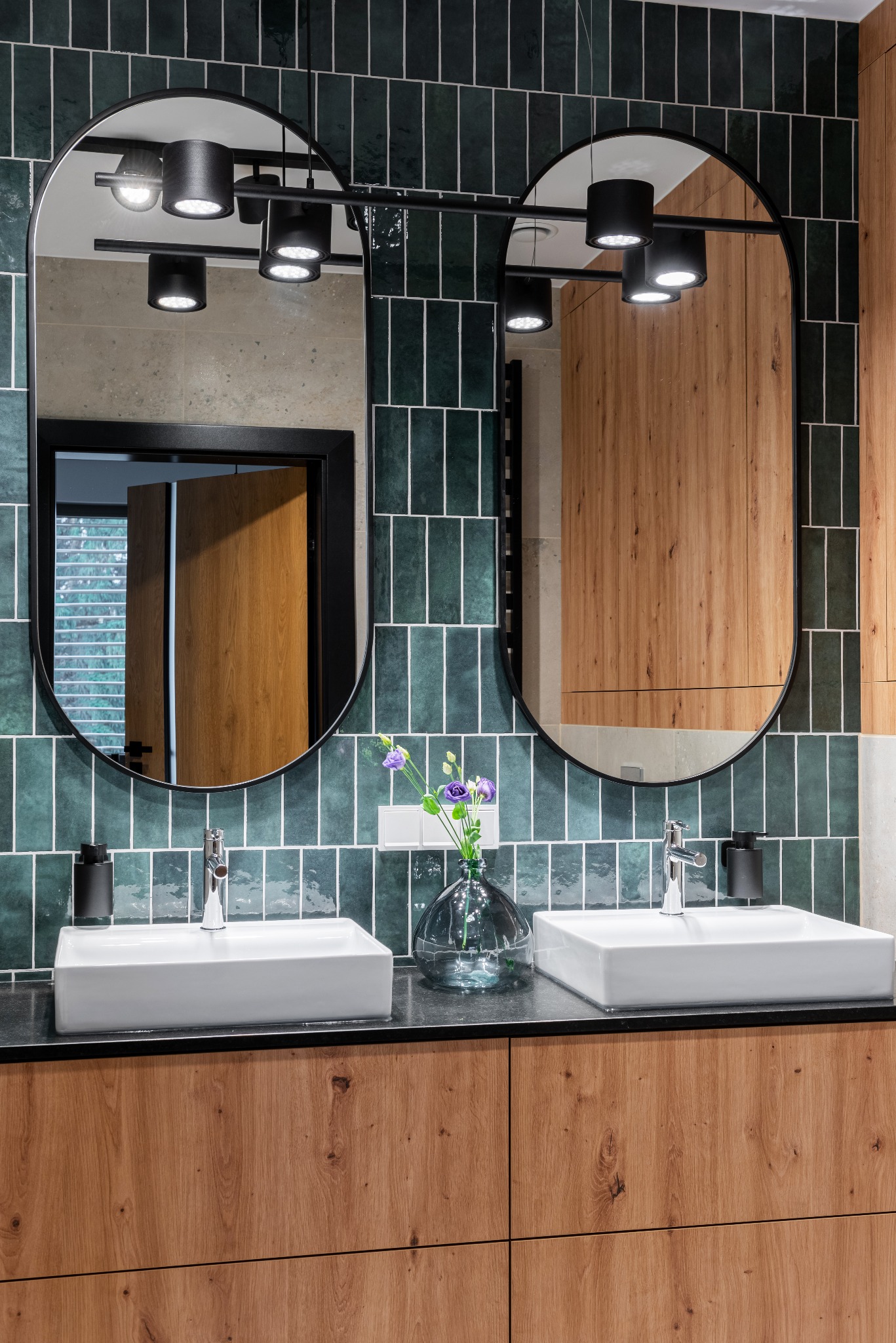 Nowoczesna łazienka z dwoma umywalkami nablatowymi, lustrami w czarnych ramach i ciemnozieloną glazurą ułożoną pionowo, oświetlenie punktowe na szynie.