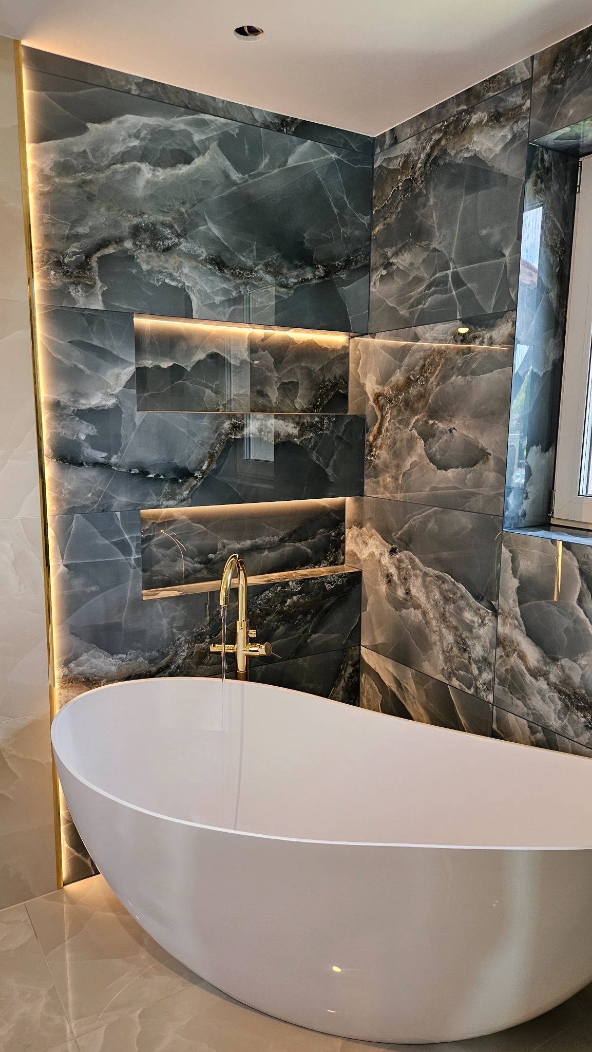 Nowoczesna łazienka z wolnostojącą białą wanną, złota bateria, ściany wyłożone płytkami imitującymi marmur z podświetleniem LED.