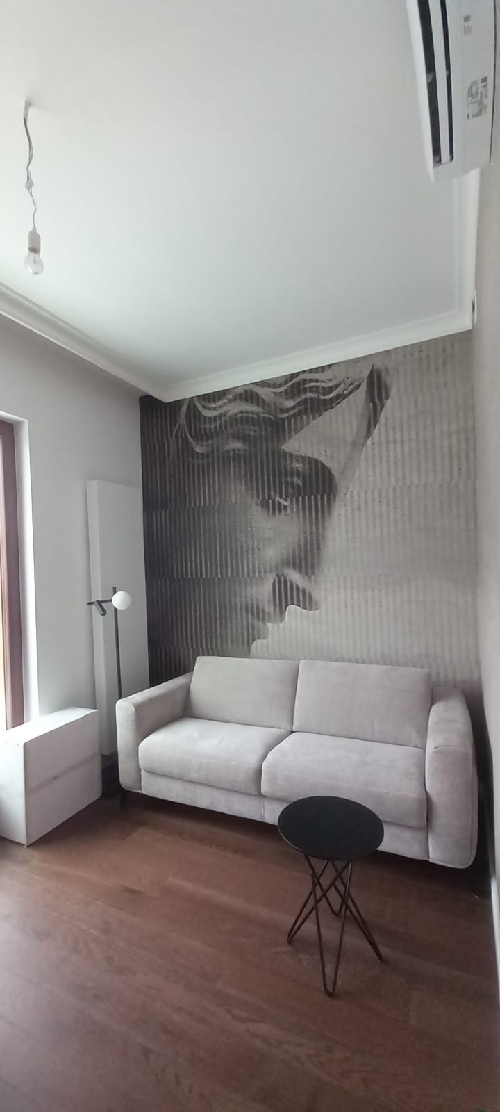 Jasnoszara sofa z dwoma poduszkami na tle ściany z fototapetą przedstawiającą twarz, obok czarny stolik kawowy na metalowych nogach i lampa podłogowa.