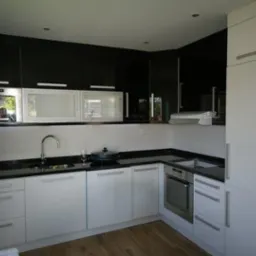 Nowoczesna kuchnia w bieli i czerni z blatem z czarnego kamienia. Uwagę przykuwają błyszczące fronty szafek i drewniana podłoga. Widoczny zlew, bateria i płyta grzewcza.