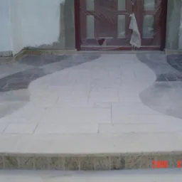 Wykończony taras kamienny przed wejściem do domu z dekoracyjnym wzorem z płytek w odcieniach szarości i beżu, wykończony kamiennym obrzeżem. Drzwi wejściowe zabezpieczone folią.