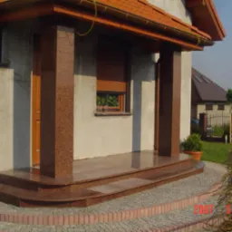 Wejście do domu z kolumnami i schodami z polerowanego kamienia w odcieniach brązu, otoczone kostką brukową. Widoczne detale wykończenia i tło z zielenią.