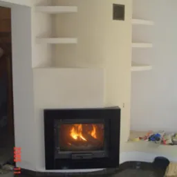 Nowoczesny kominek z czarną obudową kamienną, wbudowany w beżową ścianę z półkami. Ogień płonie w palenisku, a na podłodze widoczne ciemne płytki.