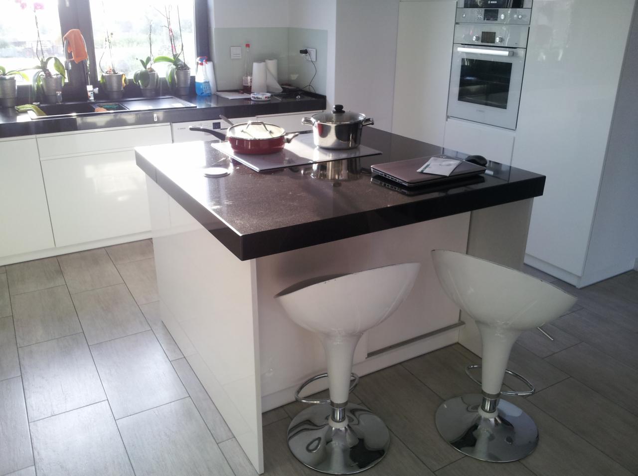 Nowoczesna kuchnia z wyspą i blatem z ciemnego kamienia, na której stoją garnki i laptop. Dwa białe krzesła barowe. Minimalistyczny design.