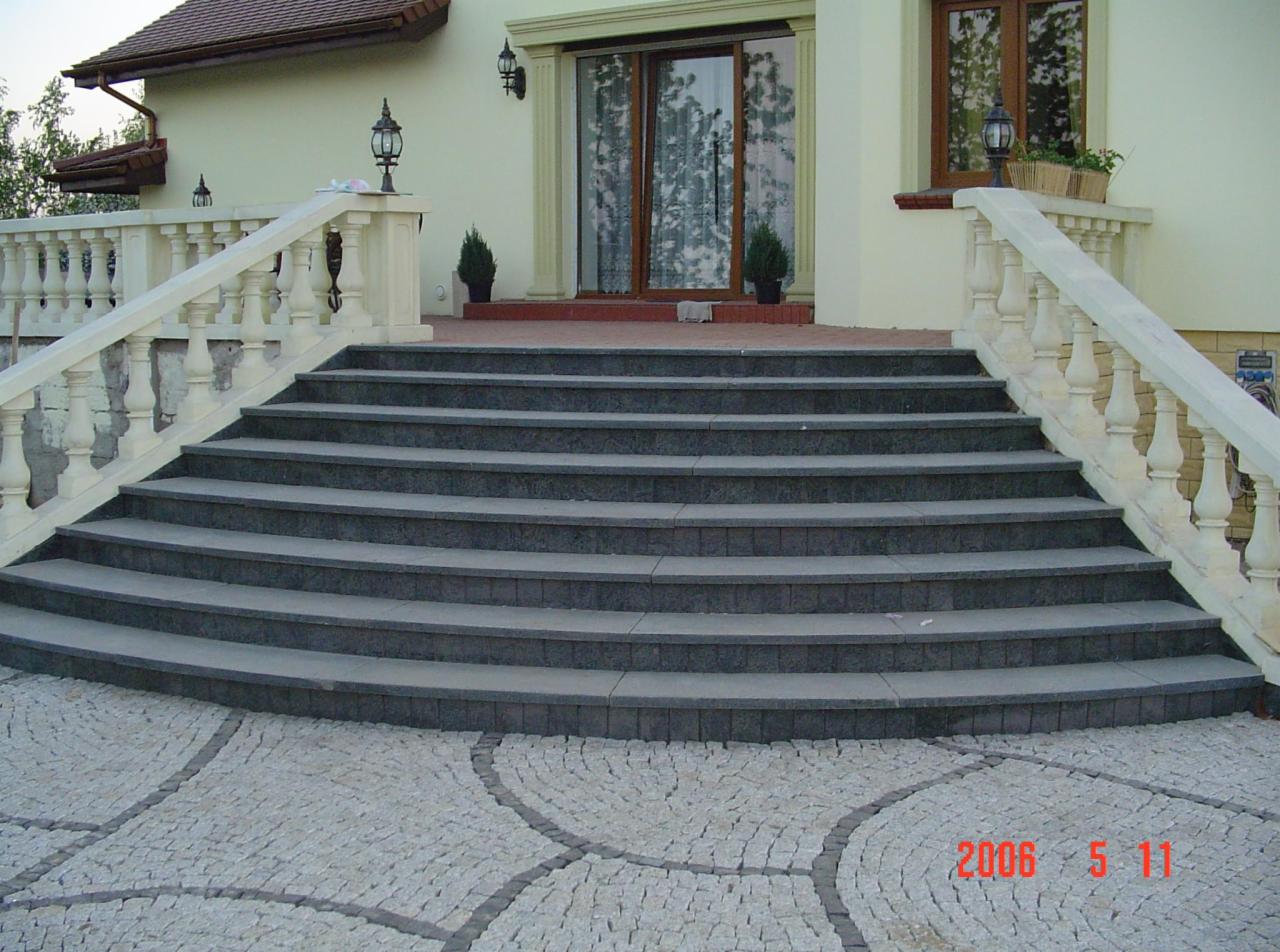 Eleganckie schody kamienne prowadzące do domu, z balustradą i kostką brukową u podstawy. Widoczne detale wykończenia i oświetlenie zewnętrzne.