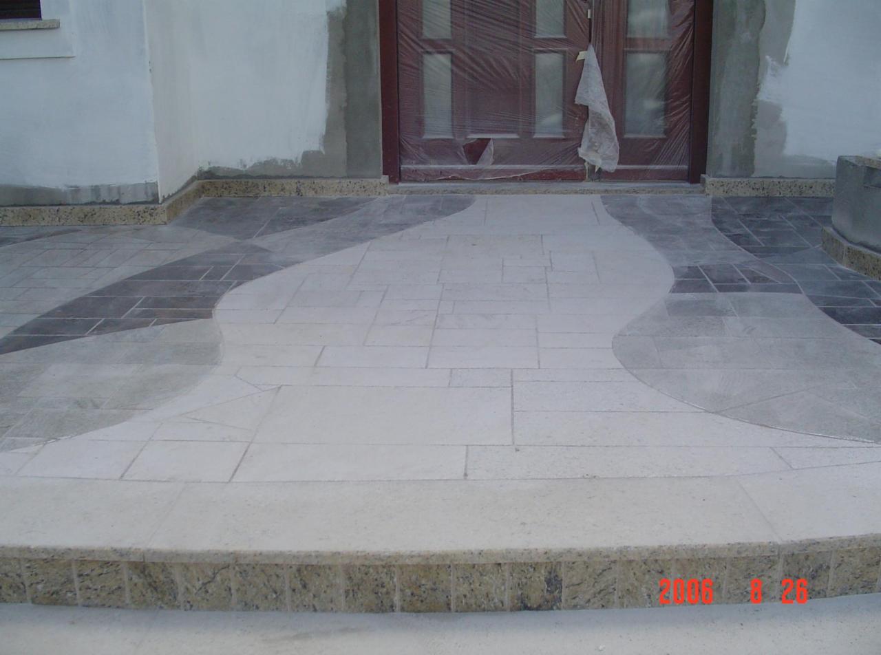 Wykończony taras kamienny przed wejściem do domu z dekoracyjnym wzorem z płytek w odcieniach szarości i beżu, wykończony kamiennym obrzeżem. Drzwi wejściowe zabezpieczone folią.