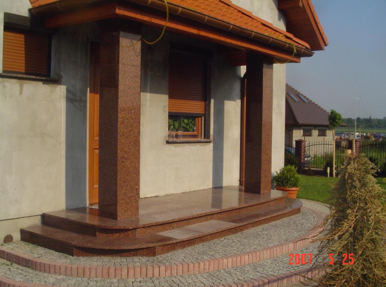 Wejście do domu z kolumnami i schodami z polerowanego kamienia w odcieniach brązu, otoczone kostką brukową. Widoczne detale wykończenia i tło z zielenią.