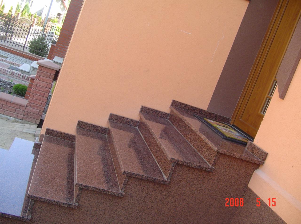 Zewnętrzne schody z granitu w odcieniach brązu, prowadzące do drzwi wejściowych. Widoczne wykończenie elewacji budynku i fragment chodnika.