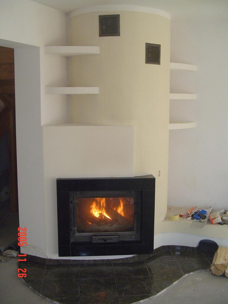 Nowoczesny kominek z czarną obudową kamienną, wbudowany w beżową ścianę z półkami. Ogień płonie w palenisku, a na podłodze widoczne ciemne płytki.