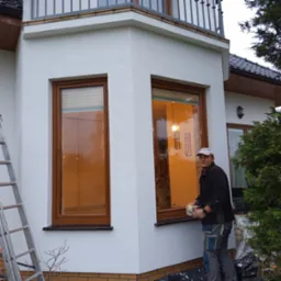 Pracownik z uśmiechem czyści elewację domu z dwoma oknami o drewnianych ramach, widoczna drabina aluminiowa i fragment zieleni