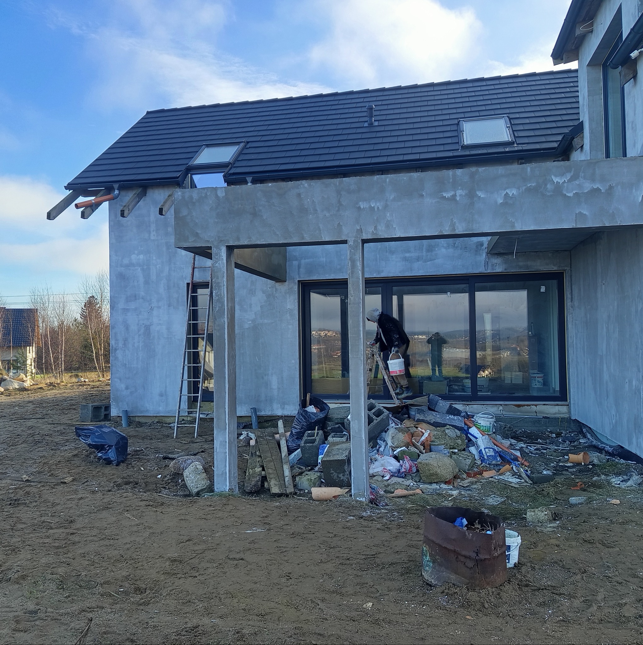 Dom w stanie surowym, z widocznymi elementami konstrukcyjnymi elewacji i stertą gruzu budowlanego na ziemi, pracownik na drabinie z wiadrem. Widoczne okna dachowe i czarna dachówka.
