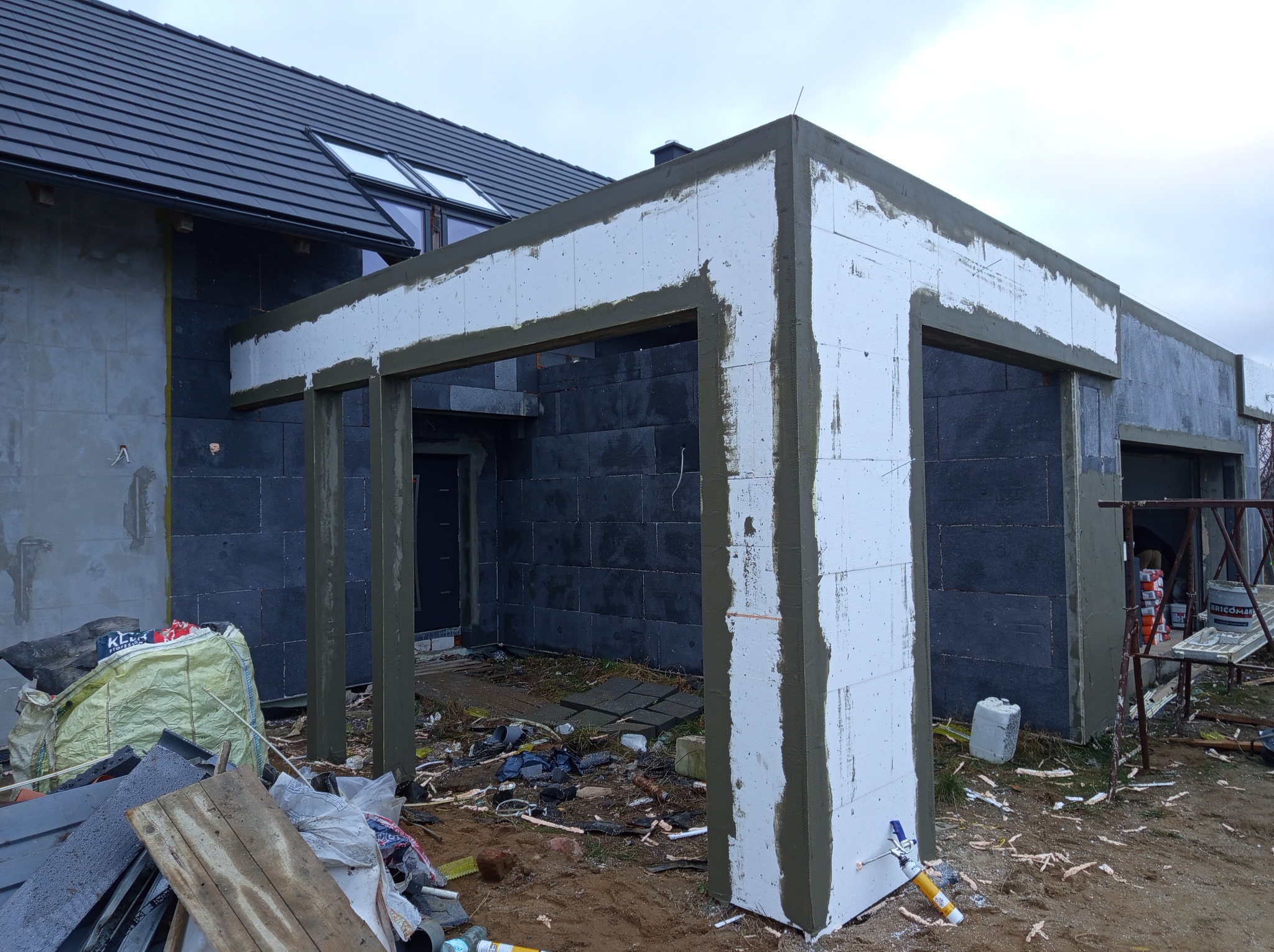 Dom w trakcie prac elewacyjnych: szara ściana z czarną okładziną, fragment ocieplenia styropianem z nałożoną warstwą kleju, widoczne narzędzia i materiały budowlane na nieuporządkowanym placu budowy.