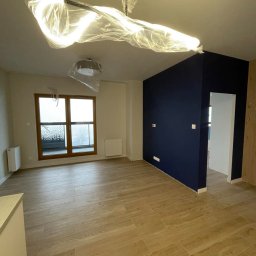 AŻ Company Arkadiusz Żurek - Wnętrze pokoju po remoncie z drewnopodobnymi panelami podłogowymi, ściana pomalowana na granatowo, okno z widokiem na balkon, lampy sufitowe zabezpieczone folią ochronną.