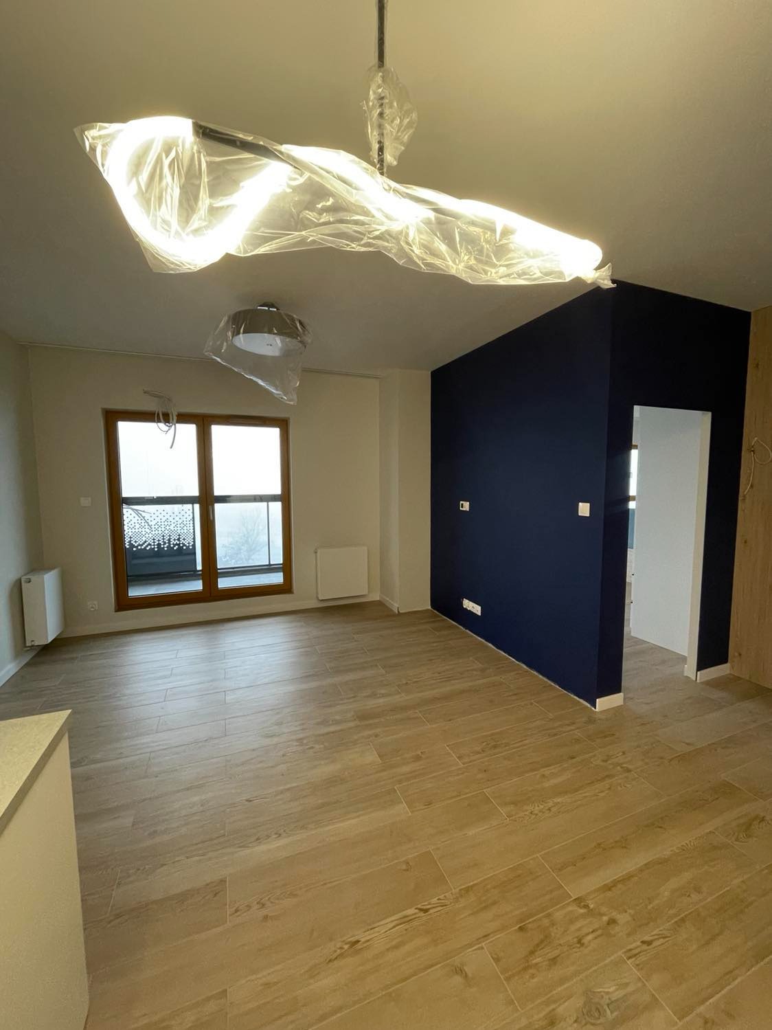 Wnętrze pokoju po remoncie z drewnopodobnymi panelami podłogowymi, ściana pomalowana na granatowo, okno z widokiem na balkon, lampy sufitowe zabezpieczone folią ochronną.