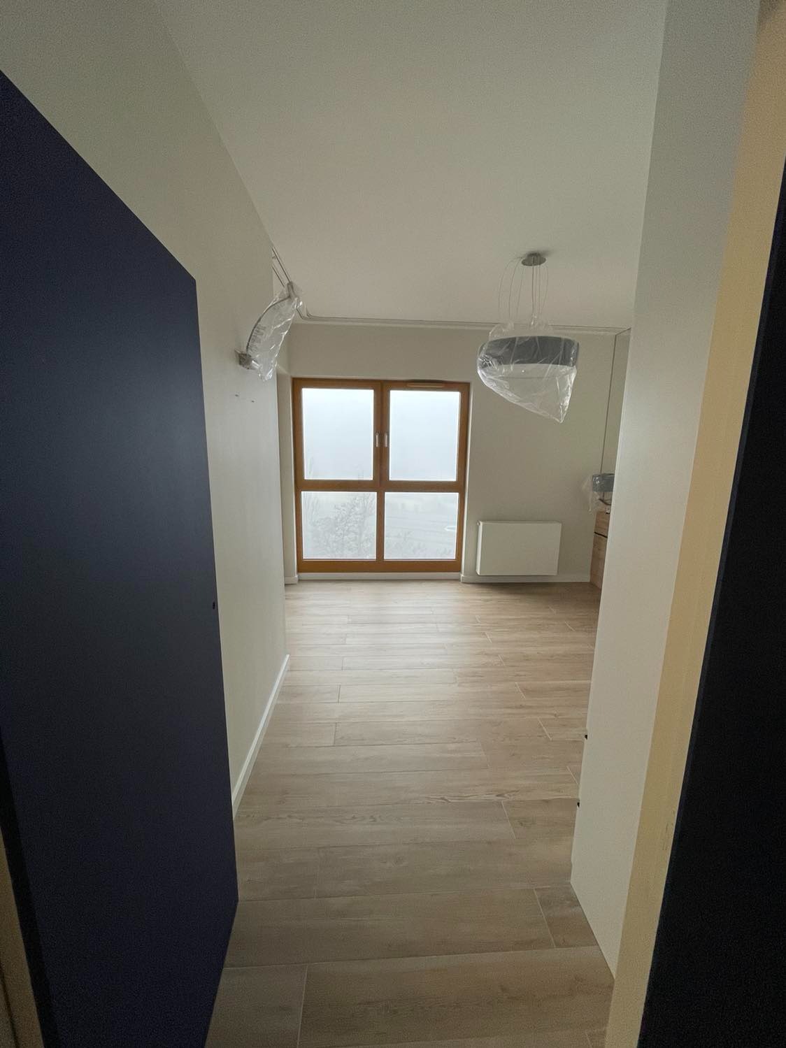 Wnętrze mieszkania po remoncie z drewnopodobną podłogą z paneli, oknem z drewnianą ramą i lampą w folii ochronnej, widok zza otwartych, granatowych drzwi.