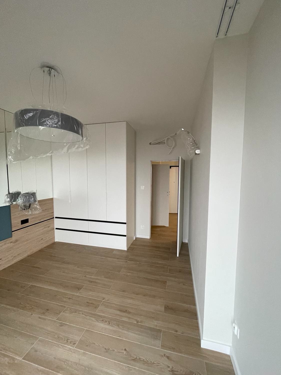 Nowoczesne wnętrze pokoju z drewnopodobną podłogą, białą szafą z czarnymi akcentami i lampą pod sufitem jeszcze w folii ochronnej, widok na otwarte drzwi do kolejnego pomieszczenia.