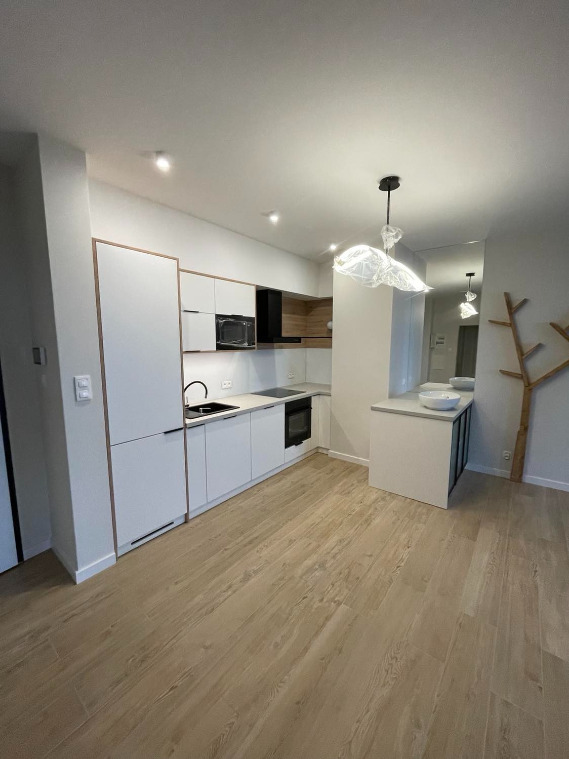 Nowoczesna, jasna kuchnia z białymi szafkami z drewnianymi obramowaniami, czarnym zlewem i płytą indukcyjną, oświetlona designerską lampą wiszącą i wykończona panelami podłogowymi w odcieniach drewna.