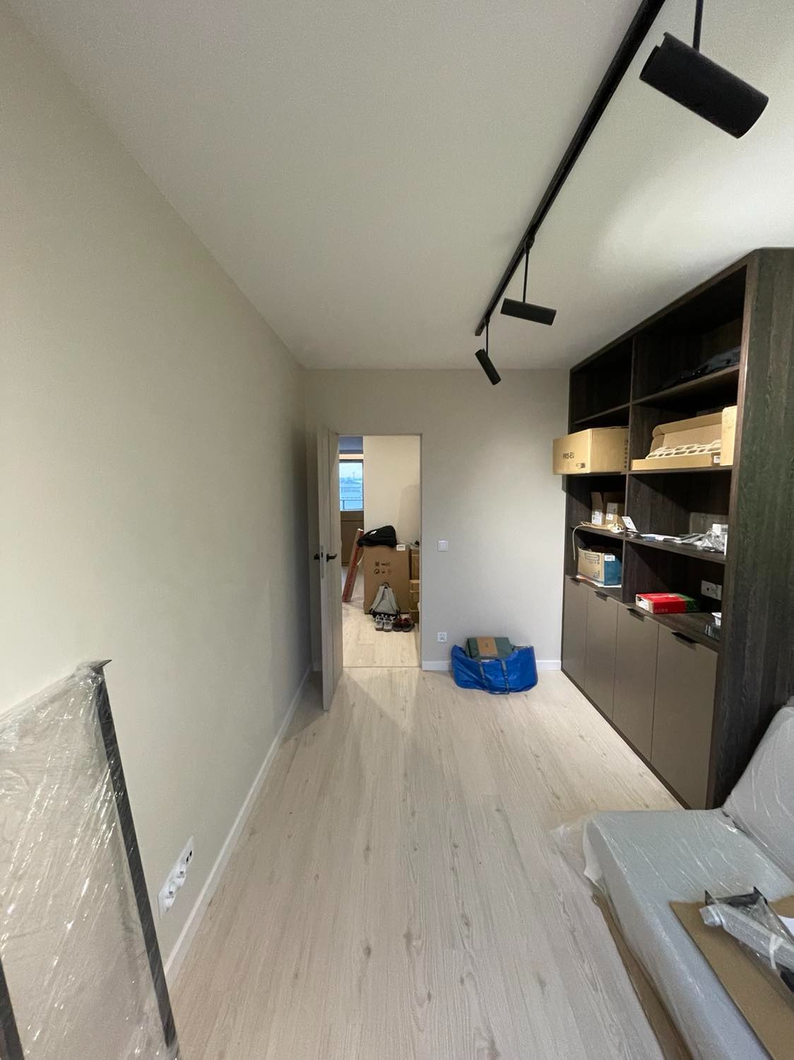 Jasne panele podłogowe w pokoju po remoncie, widoczna zabudowa meblowa w ciemnym kolorze drewna i lampa na szynie, w tle otwarte drzwi do kolejnego pomieszczenia z kartonami.