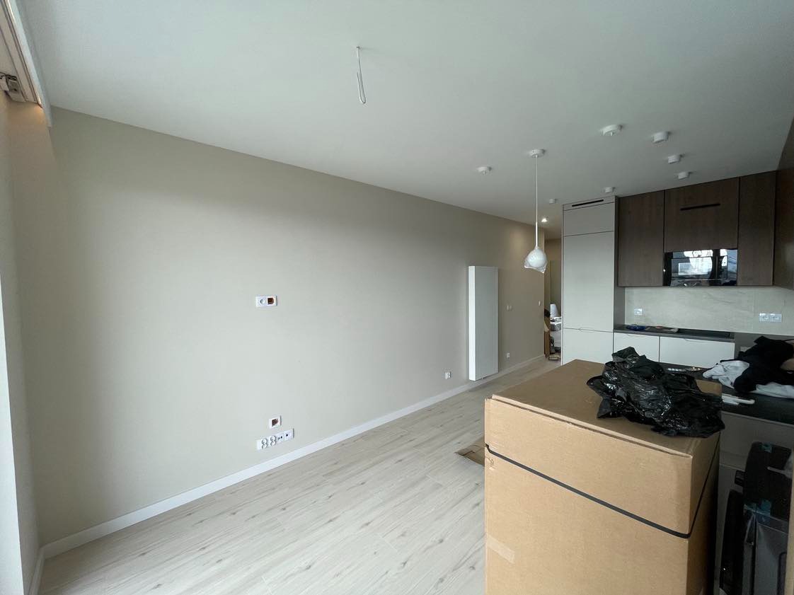 Wnętrze mieszkania w trakcie wykańczania, ściany pomalowane na beżowo, widoczne gniazdka elektryczne, jasne panele podłogowe, zabudowa kuchenna w tle, kartony z materiałami na pierwszym planie.