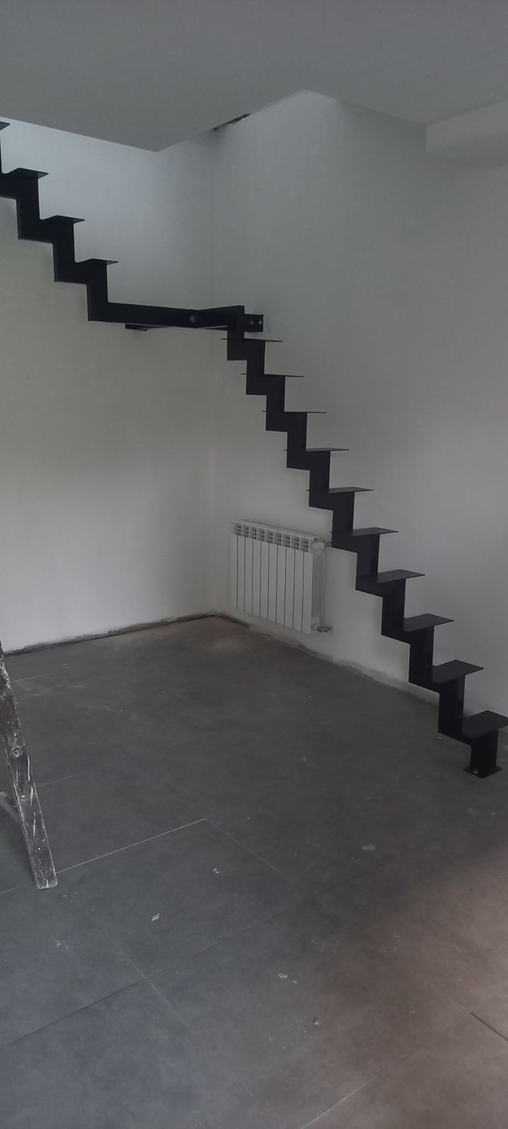 Wewnętrzne schody wspornikowe w kolorze czarnym, zamontowane przy białej ścianie, widoczny grzejnik i fragment drabiny malarskiej.