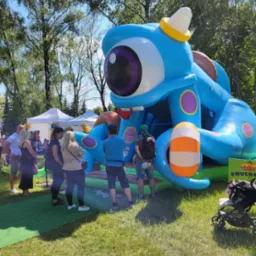 Niebieski dmuchany zamek w kształcie potwora z jednym okiem na trawie, otoczony przez ludzi i wózki dziecięce, w tle drzewa i białe namioty.