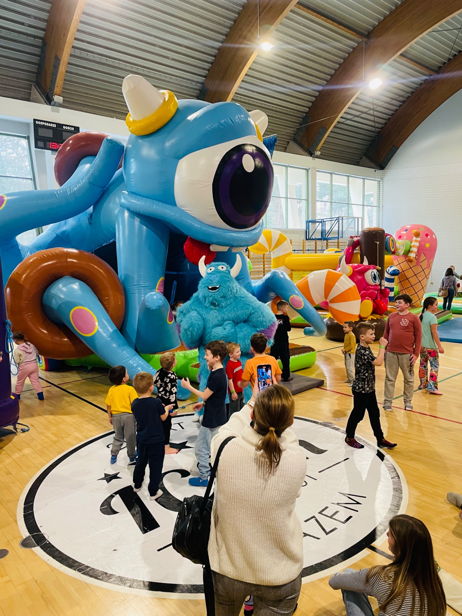 Dzieci bawią się na dmuchanym placu zabaw w hali sportowej. W tle kolorowe dmuchańce w kształcie potworów i słodyczy. Osoba w kostiumie potwora wita dzieci.