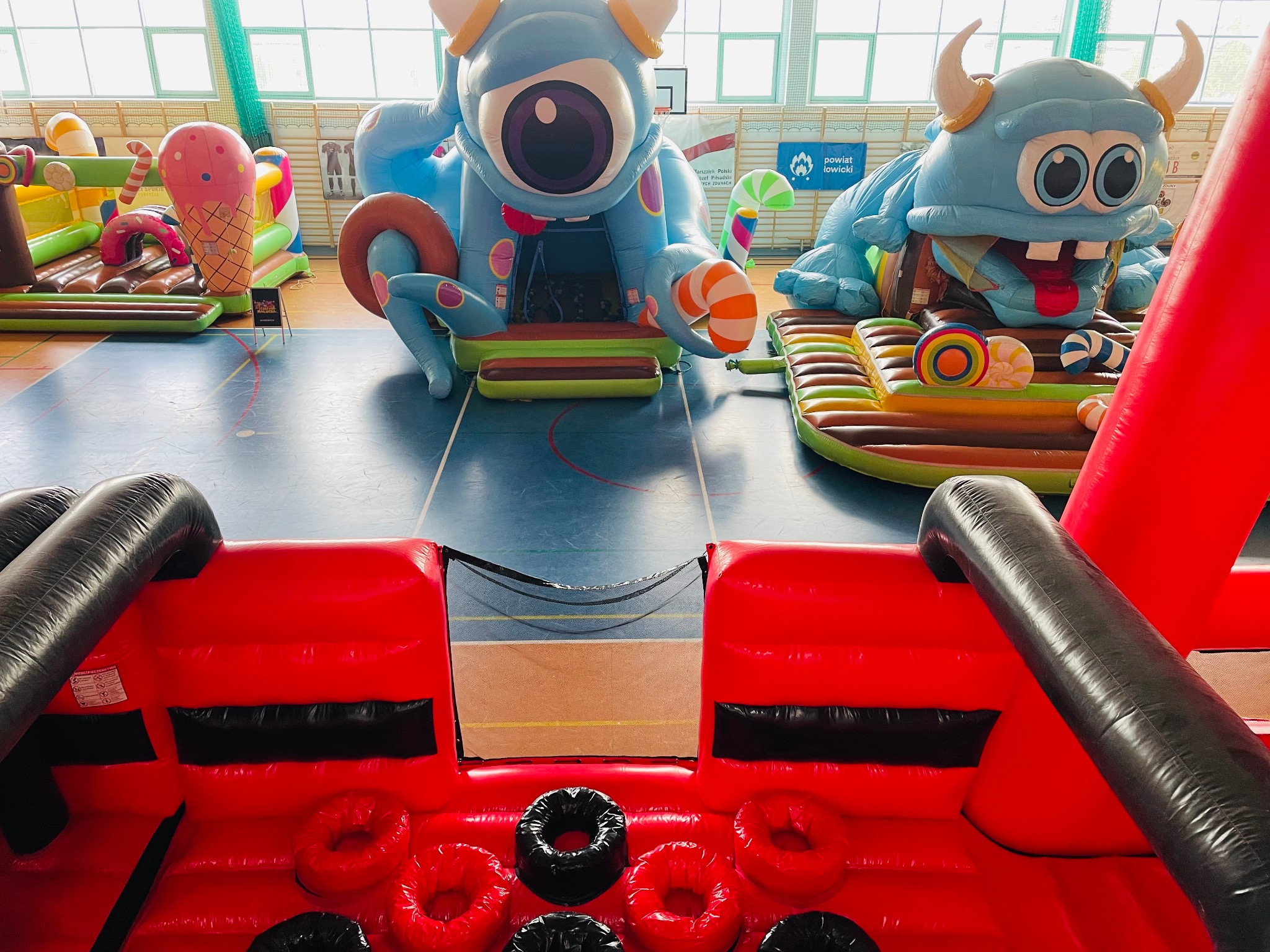 Kolorowy park dmuchany w hali sportowej, z motywami potworów i słodyczy. Widoczne elementy toru przeszkód i zjeżdżalni. Atrakcja dla dzieci na imprezie.